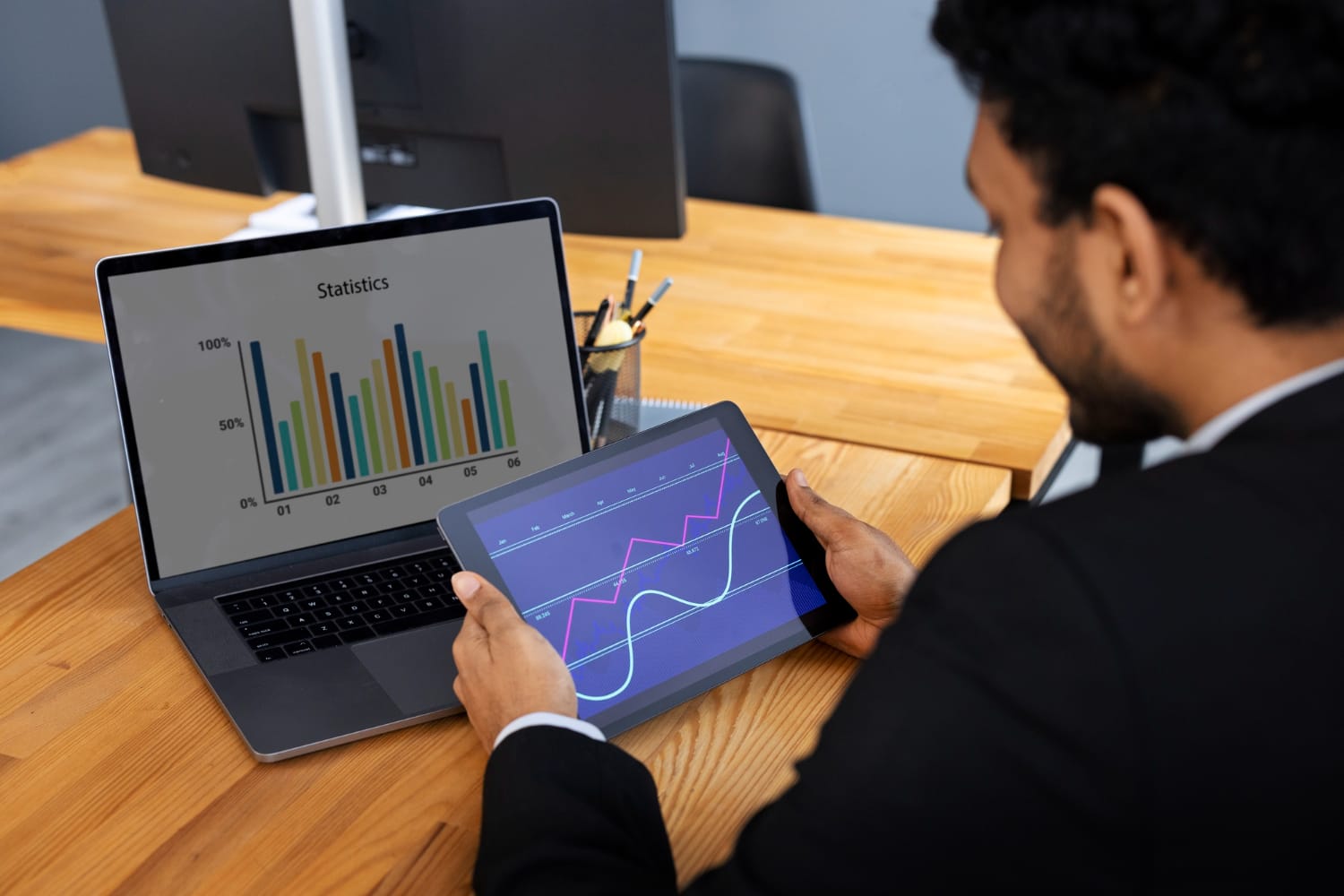 A businessman in a black suit is analyzing financial charts on a tablet while sitting at a wooden desk. A laptop in front of him displays a bar graph labeled "Statistics." The workspace includes a pen holder with pens and pencils, and a computer monitor in the background. The graphs on both screens indicate financial data analysis.