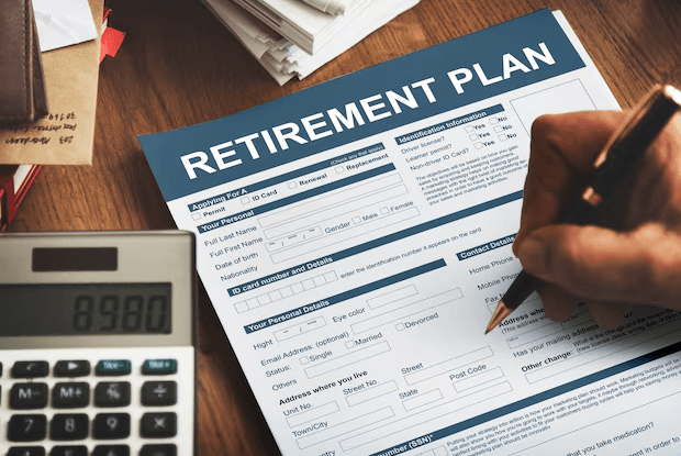 A close-up image of a person filling out a "Retirement Plan" application form with a pen. The form includes fields for personal details, identification information, and contact details. A calculator displaying "8980" and a stack of documents are placed beside the form, indicating financial planning.