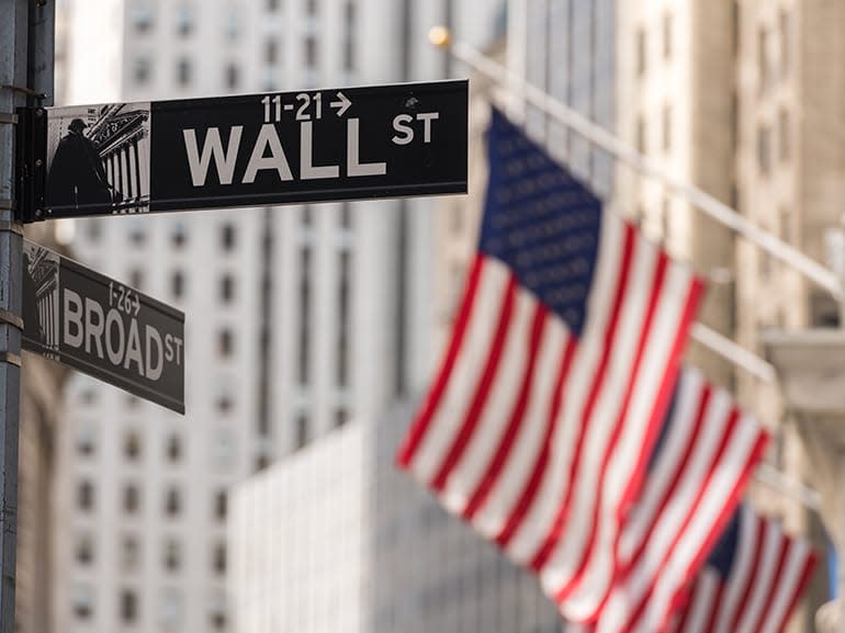 A street sign for Wall Street and Broad Street in New York City, with tall financial buildings in the background and multiple American flags hanging from poles, symbolizing the financial power of Wall Street.